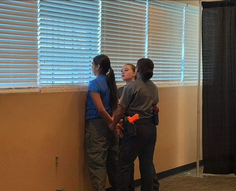 Three people standing in front of a window with blue blinds in an indoor room with tan walls
