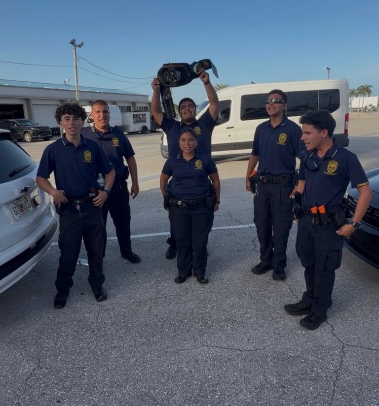 Group of six law enforcement officers in dark uniforms standing in a parking lot, one holding up a small dog above their heads