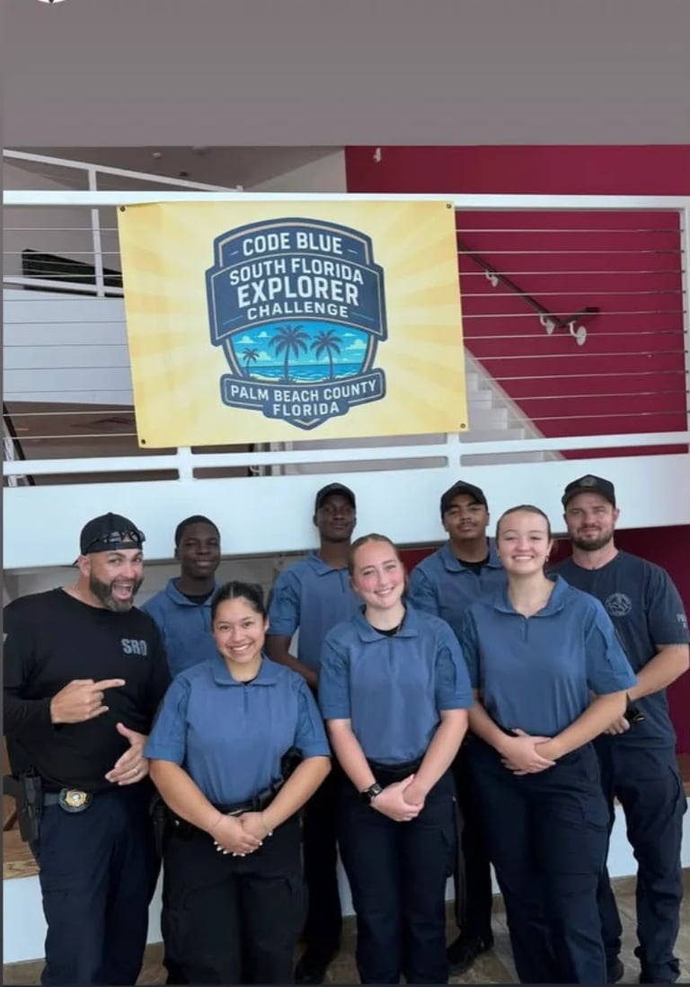 Team of eight volunteers in blue shirts posing together indoors under a Code Blue South Florida Explorer Challenge banner